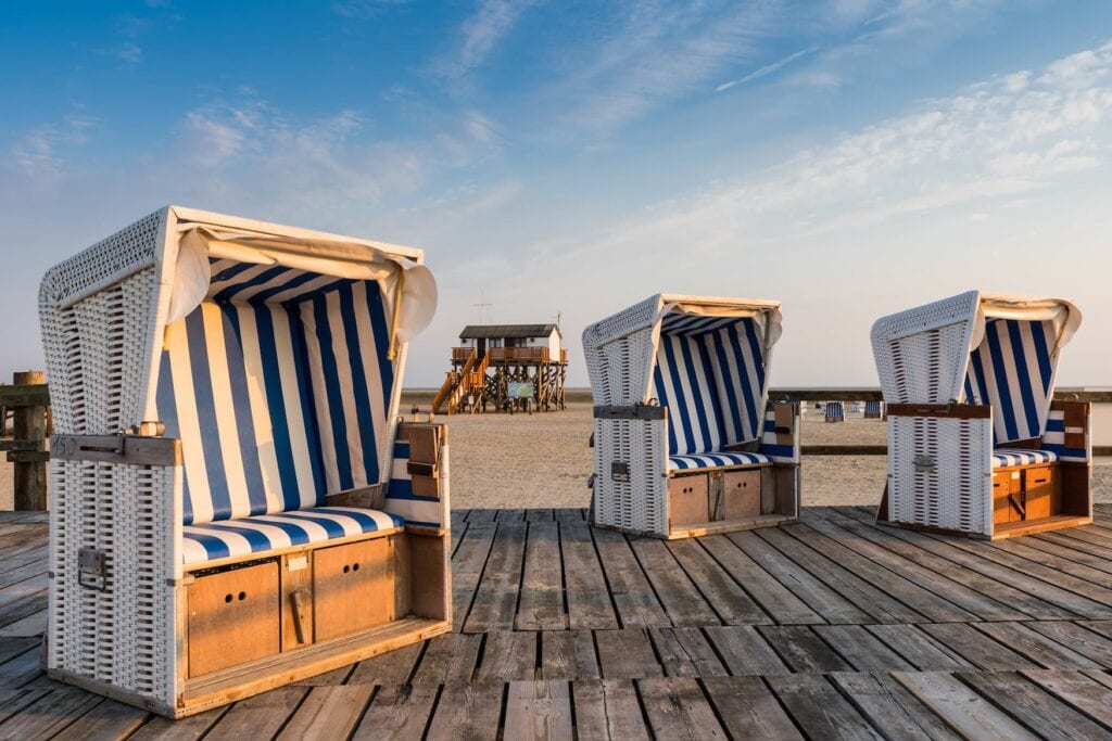 Three beach chairs on the beach at St. Peter-Ording in Schleswig-Holstein. Behind them is a small house on stilts, far in the distance the sea can be seen