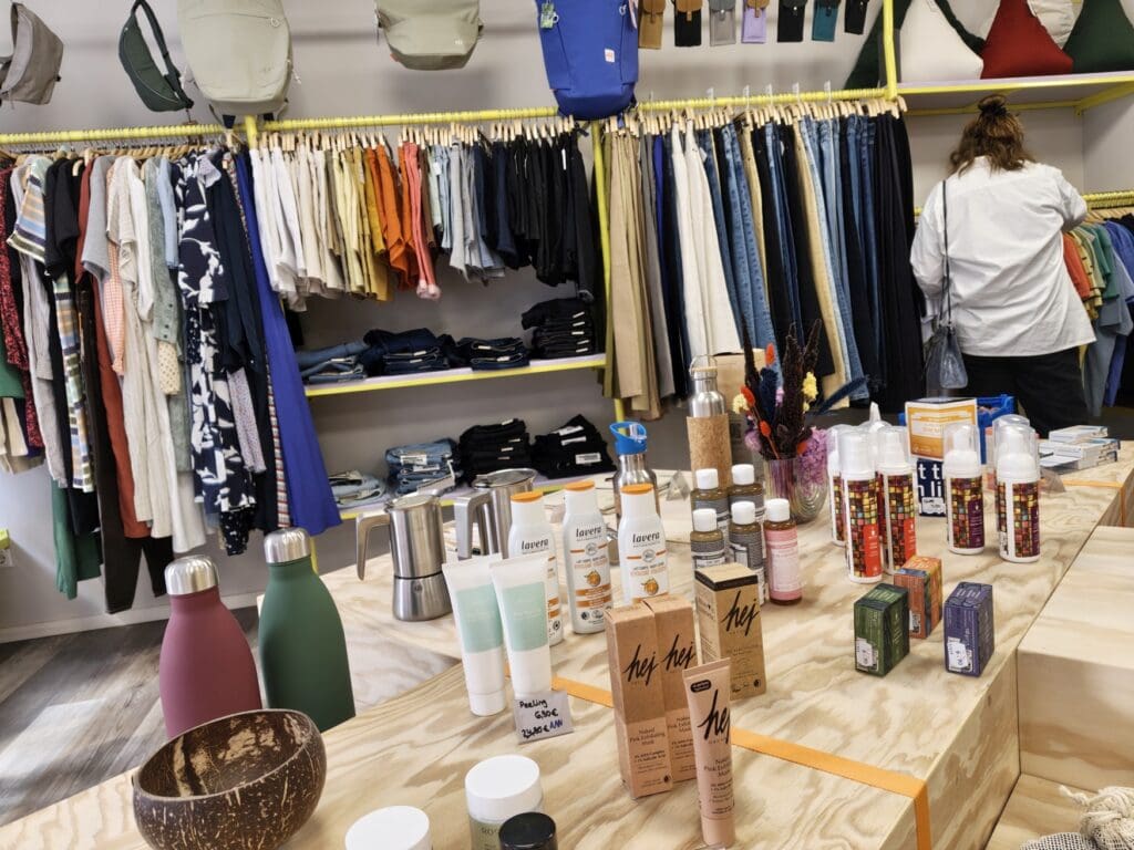 Auslage in einem Fair-Fashion-Geschäft; auf einem hölzernen Podest stehen unterschiedliche Produkte, unter anderem metallene Trinkflaschen, einige Naturkosmetikprodukte und Espressokocher. Im Hintergrund eine lange Kleiderstange mit Regalböden mit Kleidern, Röcken, Hosen und Shirts, darüber eine Reihe Taschen und Rucksäcke; rechts stöbert eine Kundin durch einige der Kleidungsstücke, man sieht ihr Gesicht nicht