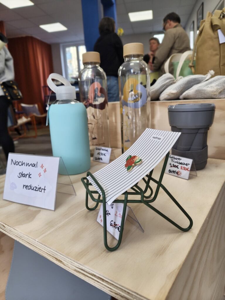 Auslage in einem Fair-Fashion-Geschäft: Auf einem Miniatur-Legestuhl ist ein Ansteck-Pin in Form einer Rose drapiert, dahinter verschiedene Mehrwegbecher- und Flaschen. Alle Produkte sind auf kleinen Schildern mit Preisen gekennzeichnet. Im Hintergrund sind unscharf einige Kunden zu erkennen