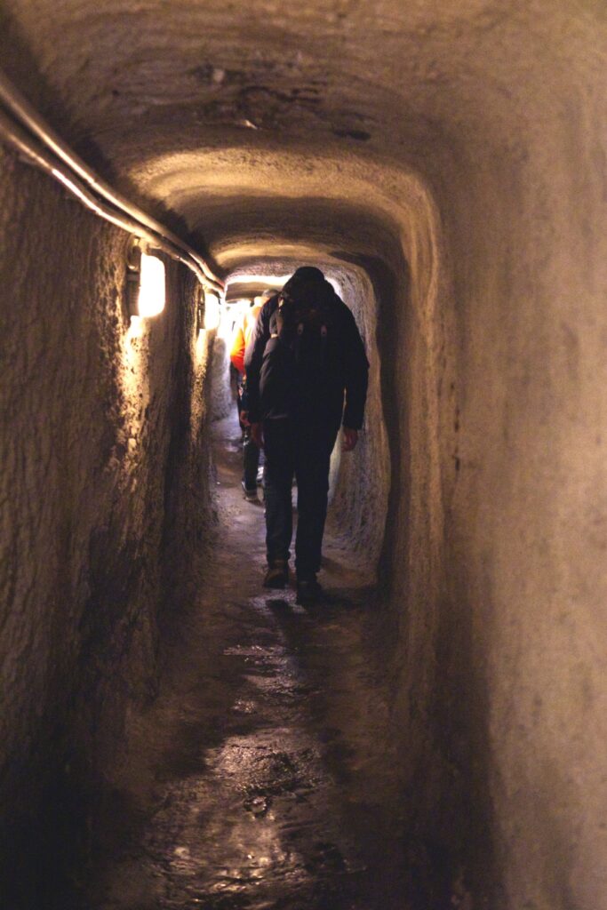 Ein niedriger, enger, unterirdischer Gang; links an der Wand einige Lampen, ein Mann bewegt sich geduckt von der Kamera weg durch den Gang