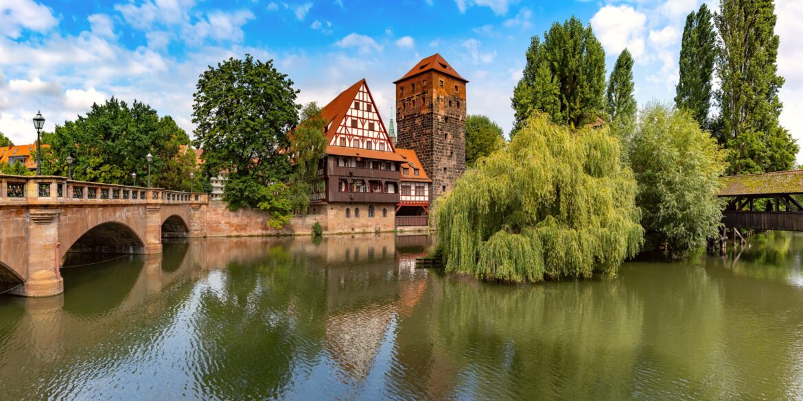 Ein breiter Fluss, von links nach rechts. Links wird der Fluss von einer Bundsandsteinbrücke mit drei Bögen überspannt; gegenüber des Betrachters eine breiter Kaimauer, auf der mittig ein mehrgeschossiges Fachwerkhaus und daneben ein eckiger Steinturm stehen. Rechts folgt neben einer großen Trauerweide eine hölzerne Brücke mit Dach.