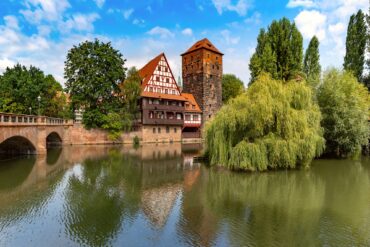 Ein breiter Fluss, von links nach rechts. Links wird der Fluss von einer Bundsandsteinbrücke mit drei Bögen überspannt; gegenüber des Betrachters eine breiter Kaimauer, auf der mittig ein mehrgeschossiges Fachwerkhaus und daneben ein eckiger Steinturm stehen. Rechts folgt neben einer großen Trauerweide eine hölzerne Brücke mit Dach.