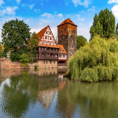 Ein breiter Fluss, von links nach rechts. Links wird der Fluss von einer Bundsandsteinbrücke mit drei Bögen überspannt; gegenüber des Betrachters eine breiter Kaimauer, auf der mittig ein mehrgeschossiges Fachwerkhaus und daneben ein eckiger Steinturm stehen. Rechts folgt neben einer großen Trauerweide eine hölzerne Brücke mit Dach.