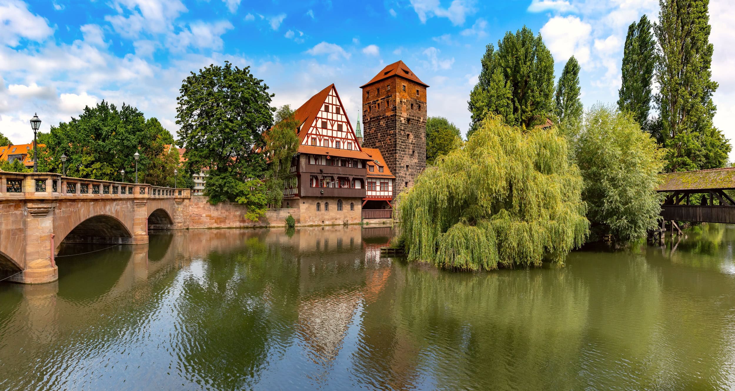 Ein breiter Fluss, von links nach rechts. Links wird der Fluss von einer Bundsandsteinbrücke mit drei Bögen überspannt; gegenüber des Betrachters eine breiter Kaimauer, auf der mittig ein mehrgeschossiges Fachwerkhaus und daneben ein eckiger Steinturm stehen. Rechts folgt neben einer großen Trauerweide eine hölzerne Brücke mit Dach.
