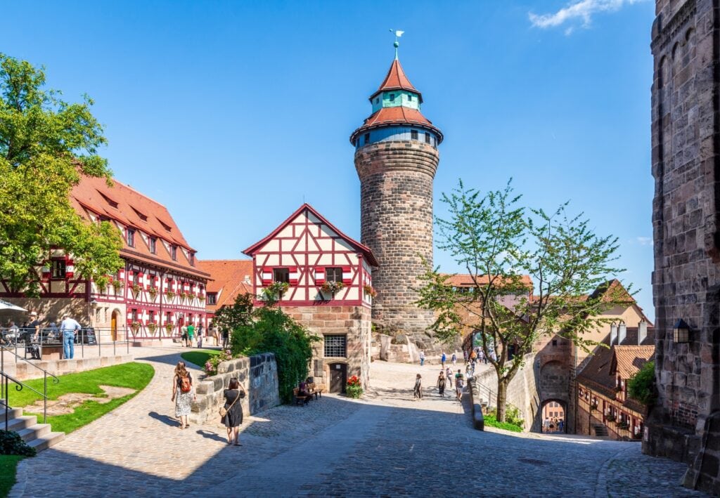 Der sonnenbeschienene Innenhof einer Burg. Ganz links ein Baum, daneben zwei Wohngebäude mit rot-weißem Fachwerk. Prominent mittig ein dicker Rundturm, der alle anderen Gebäude überragt. Rechts davon ein weiteres Gebäude, unter dem eine Rampe hinab aus dem Hof durch einen Torbogen hinausführt