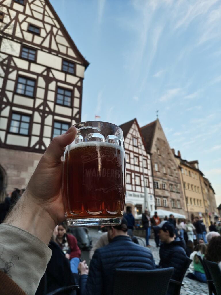 Eine Hand hält ein Bierglas mit dunklem Bier die Höhe; dahinter ein Platz mit vielen Leuten, die in kleinen Gruppen zusammensitzen, teils auf dem Boden. Hinter alledem eine Reihe mehrgeschossiger Fachwerkhäuser unter blauem Himmel