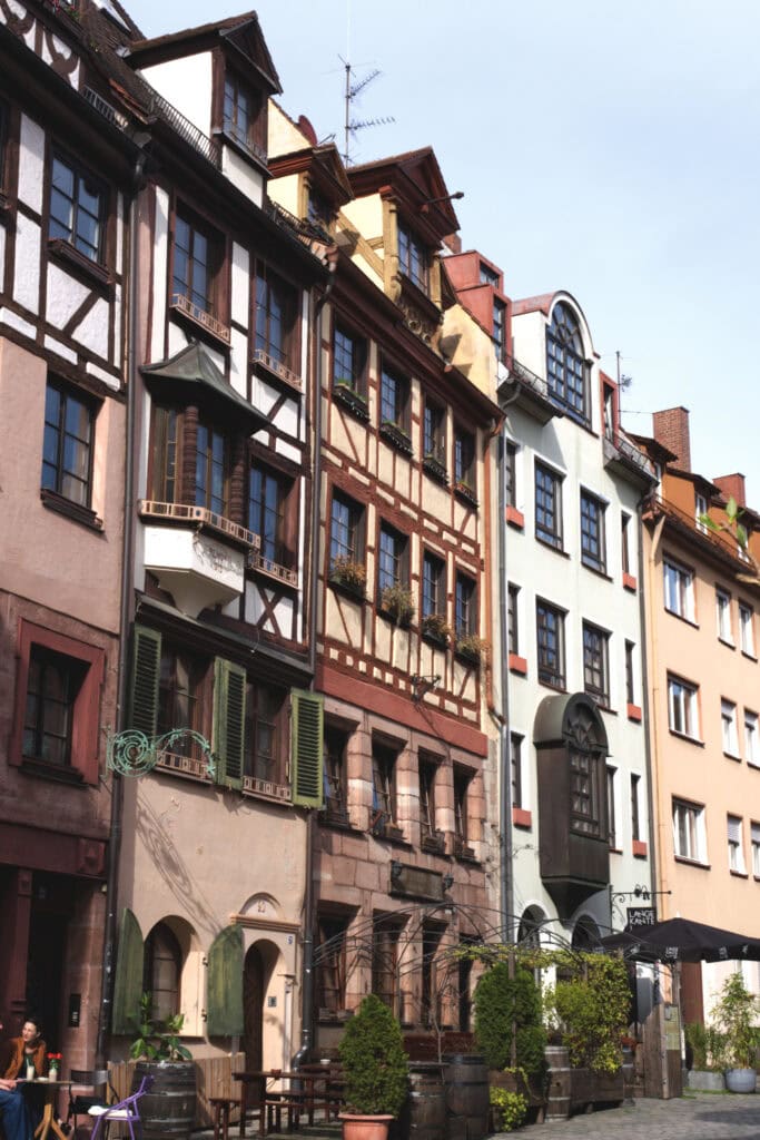 Fünf Häuser stehen dicht an dicht nebeneinander; die drei linken weisen in den oberen Stockwerken Fachwerk auf, einige Blumen sind an den Fenstern angebreacht. Die Straße vor den Häusern ist gepflastert, vor dem Haus ganz links steht ein kleiner Tisch, an dem zwei Personen in der Sonne sitzen.
