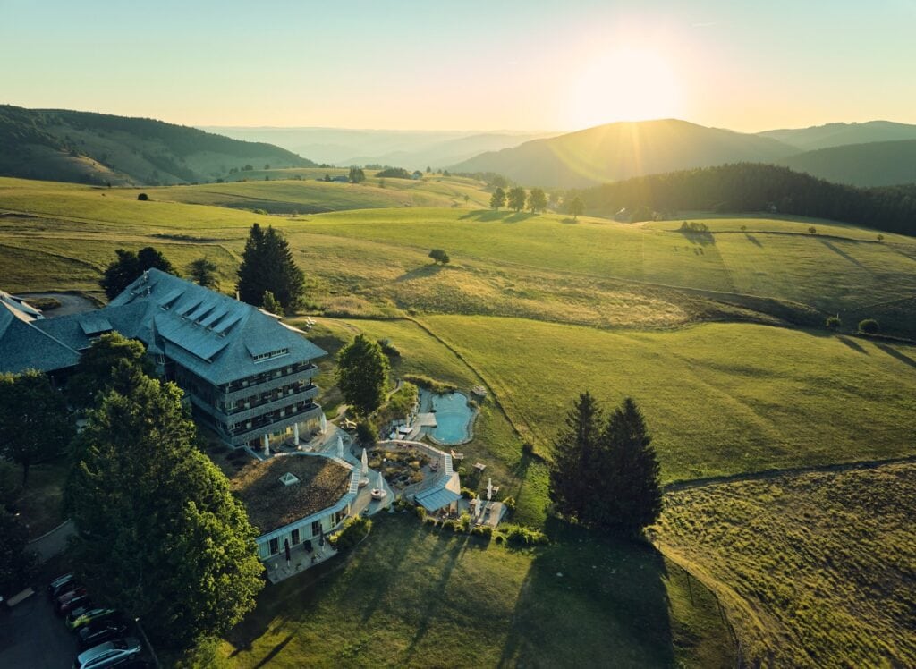 Drohnenaufnahme eines Hotels im Schwarzwald; links unten ist das mehrgeschossige Hotel in einer modernen Form der Schwarzwälder Architektur, umgeben von einigen Bäumen; dahinter erstreckt sich eine weite, in mehreren Hügeln geschwungene Wiese. Am Horizont sind im Licht der untergehenden Sonne die Silhouetten weitere bewaldete Gipfel des Schwarzwaldes zu sehen