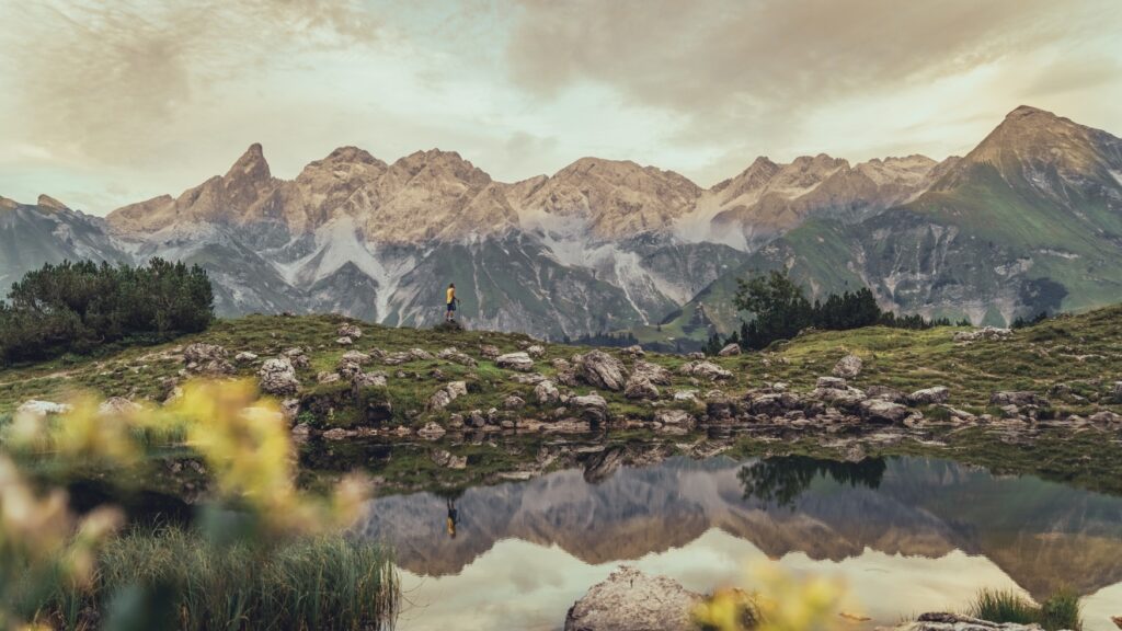 Ein Bergsee, in dem sich die Berge am Horizont spiegeln. Das Ufer ist eine von Felsen gespickte Wiese, auf einer Kammanhöhe steht eine Person in einer gelben Jacke