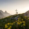 Eine Person steht neben einem Gipfelkreuz; das Kreuz steht inmitten einer Wiese mit gelben Blumen. Hinte dem Gipfel tauchen einige weitere am Horizont auf