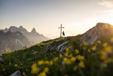 Eine Person steht neben einem Gipfelkreuz; das Kreuz steht inmitten einer Wiese mit gelben Blumen. Hinte dem Gipfel tauchen einige weitere am Horizont auf