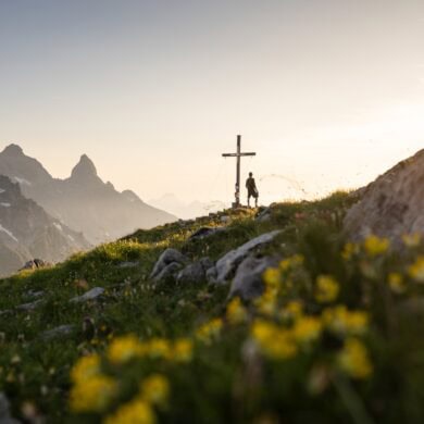 Eine Person steht neben einem Gipfelkreuz; das Kreuz steht inmitten einer Wiese mit gelben Blumen. Hinte dem Gipfel tauchen einige weitere am Horizont auf