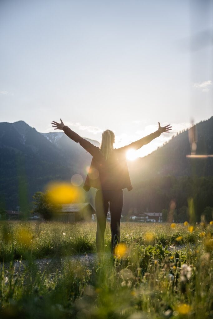 Eine Frau steht auf einer Wiese mit einigen gelben Blumen, den Rücken zum Betrachter. Sie streckt die Arme von sich weg zu beiden Seiten in die Höhe, als würde sie die Sonne, die hinter den Bergen am Horizont aufgeht, begrüßen.