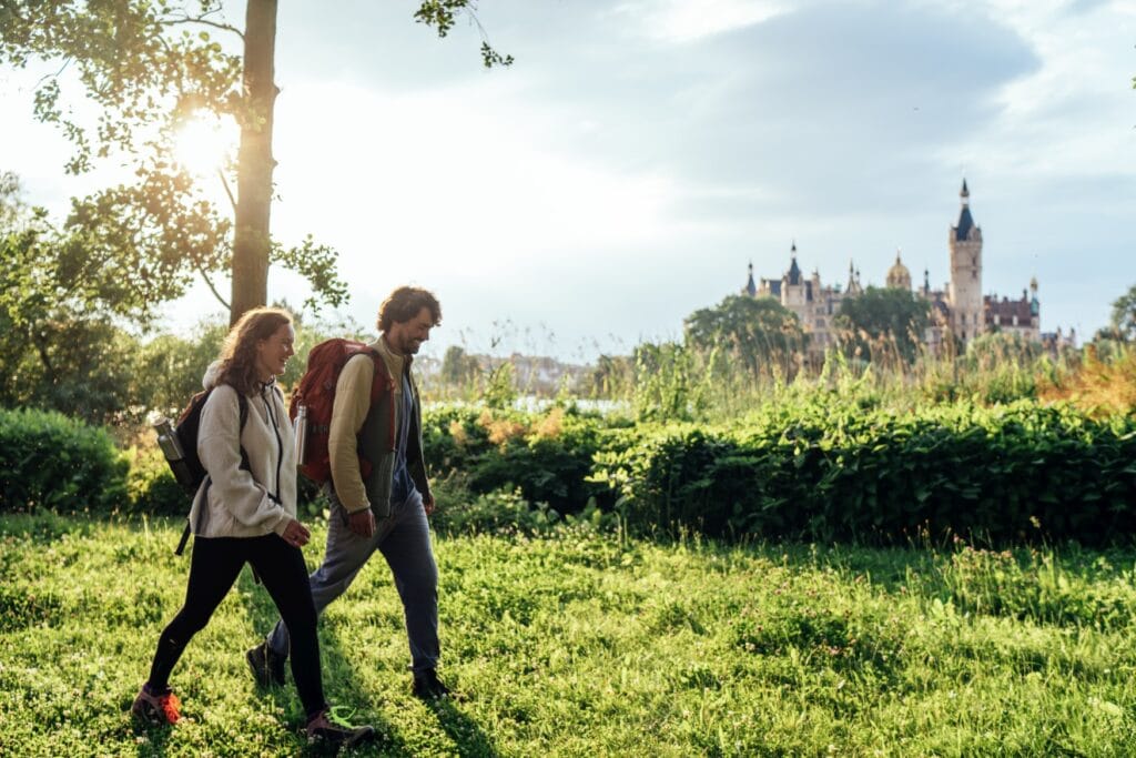 Ein Mann und eine Frau in Funktionskleidung wandern vor einer Hecke über eine grüne Wiese. Hinter der Hecke ist unscharf im Hintergrund das Schweriner Schloss zu erkennen.