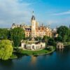 Das Schweriner Schloss aus der Luft aufgenommen, mit Blick auf den Schlossgarten im Zentrum. Die Fassade strahlt hell im Sonnenlicht, rechts von der Insel ist die Stadt zu sehen, aus der sich eine Backsteinkirche erhebt.