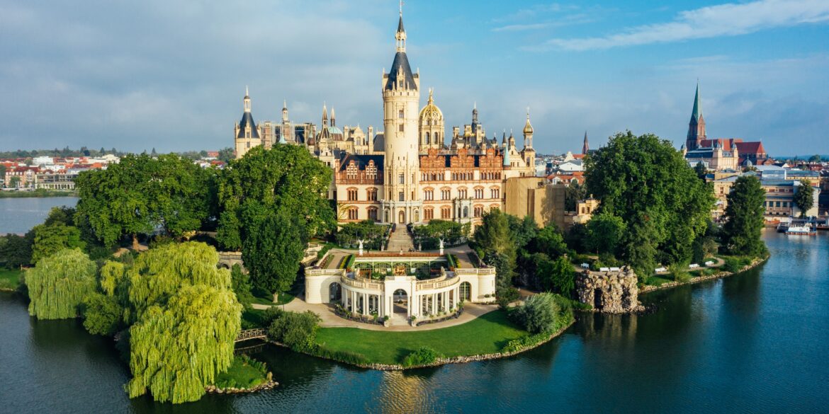 Das Schweriner Schloss aus der Luft aufgenommen, mit Blick auf den Schlossgarten im Zentrum. Die Fassade strahlt hell im Sonnenlicht, rechts von der Insel ist die Stadt zu sehen, aus der sich eine Backsteinkirche erhebt.