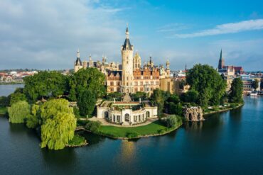 Das Schweriner Schloss aus der Luft aufgenommen, mit Blick auf den Schlossgarten im Zentrum. Die Fassade strahlt hell im Sonnenlicht, rechts von der Insel ist die Stadt zu sehen, aus der sich eine Backsteinkirche erhebt.