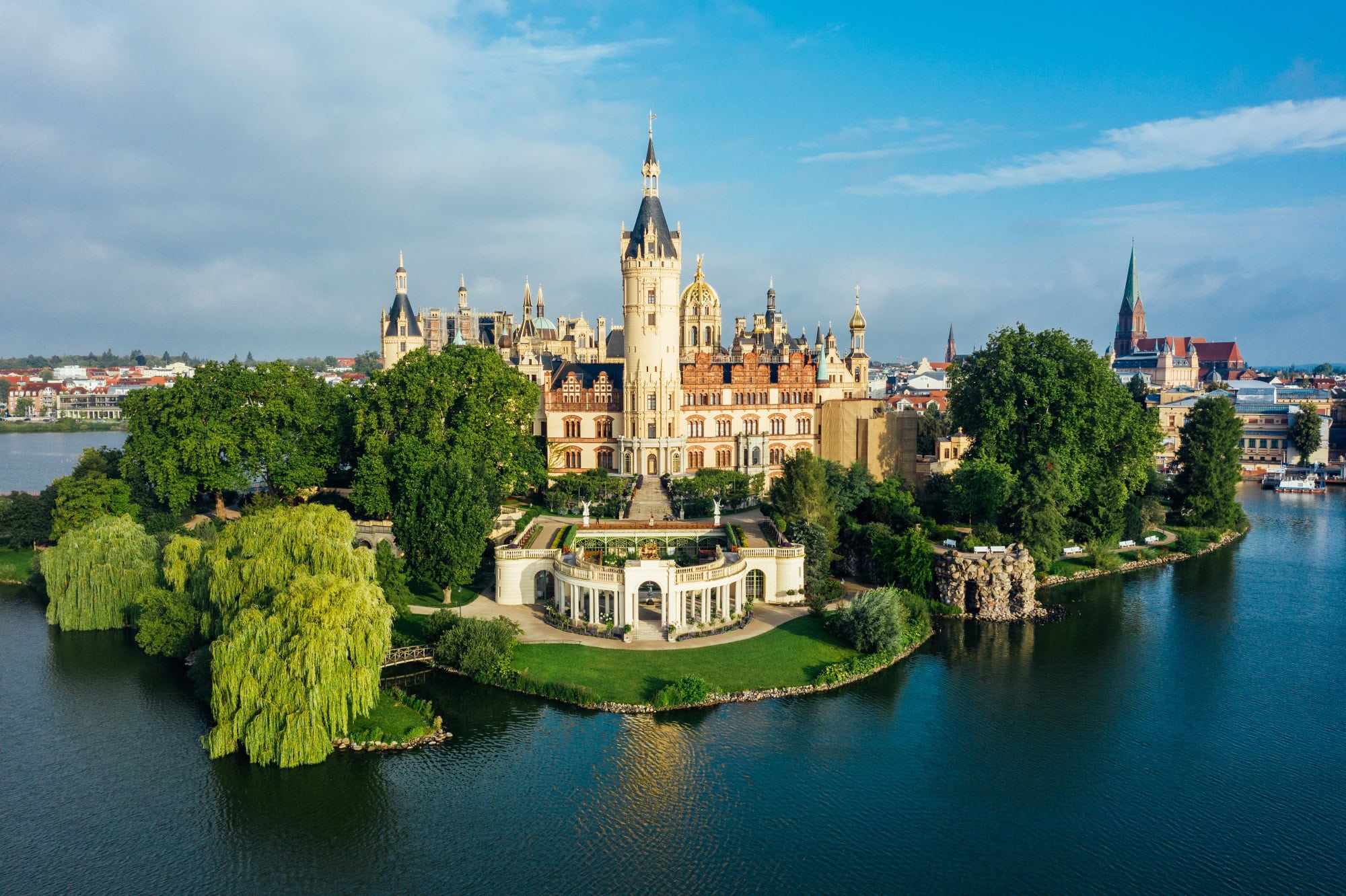 Das Schweriner Schloss aus der Luft aufgenommen, mit Blick auf den Schlossgarten im Zentrum. Die Fassade strahlt hell im Sonnenlicht, rechts von der Insel ist die Stadt zu sehen, aus der sich eine Backsteinkirche erhebt.