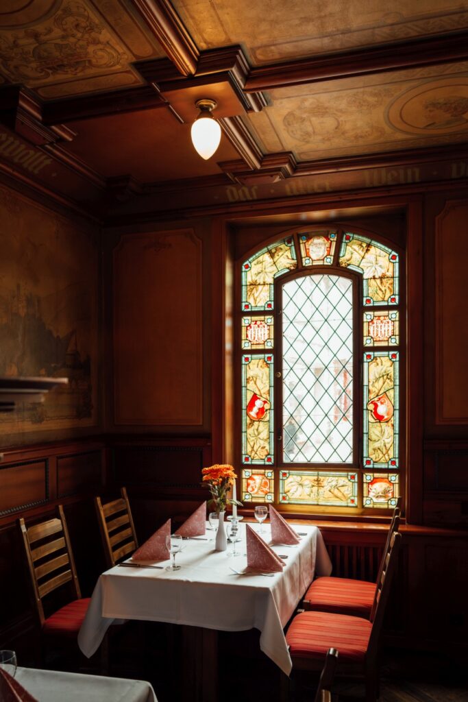 Eine Sitznische in einem historischen Gasthaus: In der Nische steht ein gedeckter Tisch mit je zwei gepolsterten Holzstühlen an den Seiten. Am Kopfende des Tisches schließt ein hohes Bleiglasfenster mit bunten Scheiben an, durch das ein gemütliches Licht fällt. Der Rest des Raums ist holzvertäfelt.