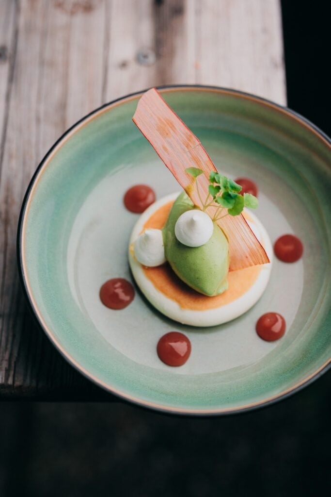 Nahufnahme eines Fine-Dining-Desserts: Eine grüne Creme (vielleicht Eis) liegt auf einem weißen Teigfladen, dazwischen eine Art orangene Oblate. Um den Fladen sind sechs Kleckse rote Soße angeordnet, alles zusammen in einem tiefen Keramikteller, der auf der Innenseite eine türkise Glasur zeigt
