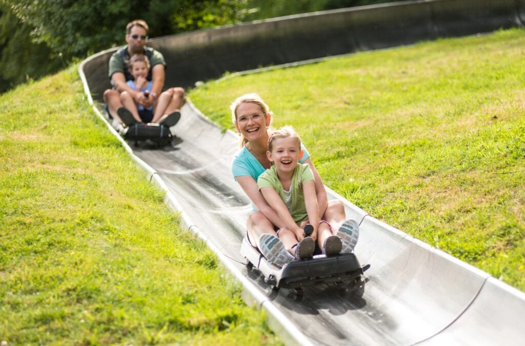 Zwei Schlitten mit je einer erwachsenen Person und einem Kind fahren auf einer metallenen Sommerrodelbahn einen Hang hinab