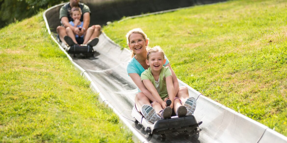 Zwei Schlitten mit je einer erwachsenen Person und einem Kind fahren auf einer metallenen Sommerrodelbahn einen Hang hinab