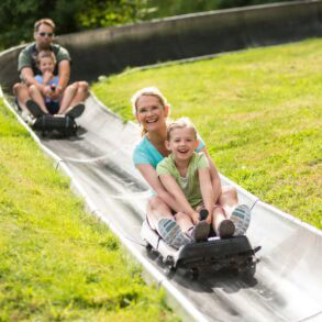 Zwei Schlitten mit je einer erwachsenen Person und einem Kind fahren auf einer metallenen Sommerrodelbahn einen Hang hinab