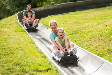 Zwei Schlitten mit je einer erwachsenen Person und einem Kind fahren auf einer metallenen Sommerrodelbahn einen Hang hinab