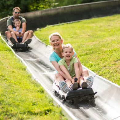 Zwei Schlitten mit je einer erwachsenen Person und einem Kind fahren auf einer metallenen Sommerrodelbahn einen Hang hinab