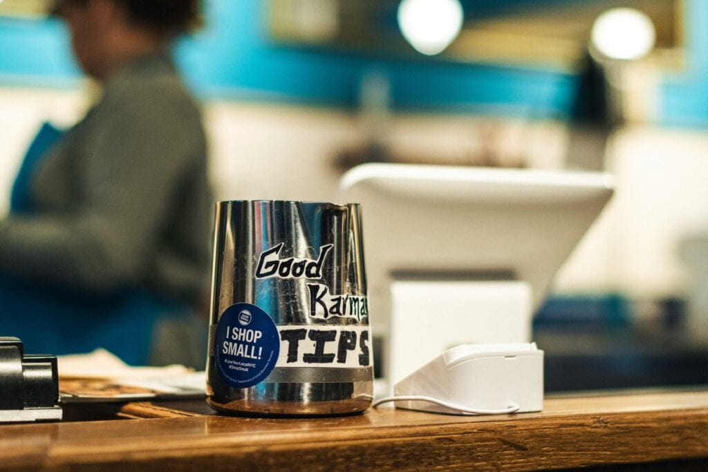 Close-up of a wooden counter in a café. On it is a milk jug with a handwritten sticker that reads “Good Karma - TIPS,” next to it a card reader and, slightly blurred in the background, an electronic cash register.