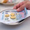 A metal tablet with a 20-Euro-bill atop of a receipt and some cents on it, grabbed by a hand