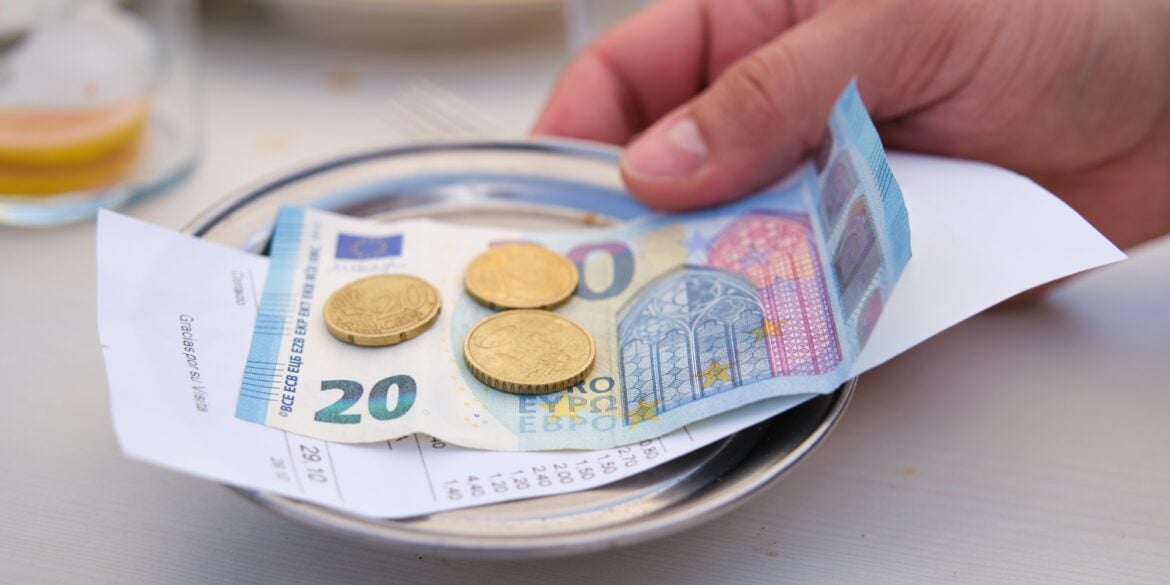 A metal tablet with a 20-Euro-bill atop of a receipt and some cents on it, grabbed by a hand