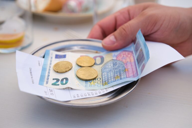 A metal tablet with a 20-Euro-bill atop of a receipt and some cents on it, grabbed by a hand
