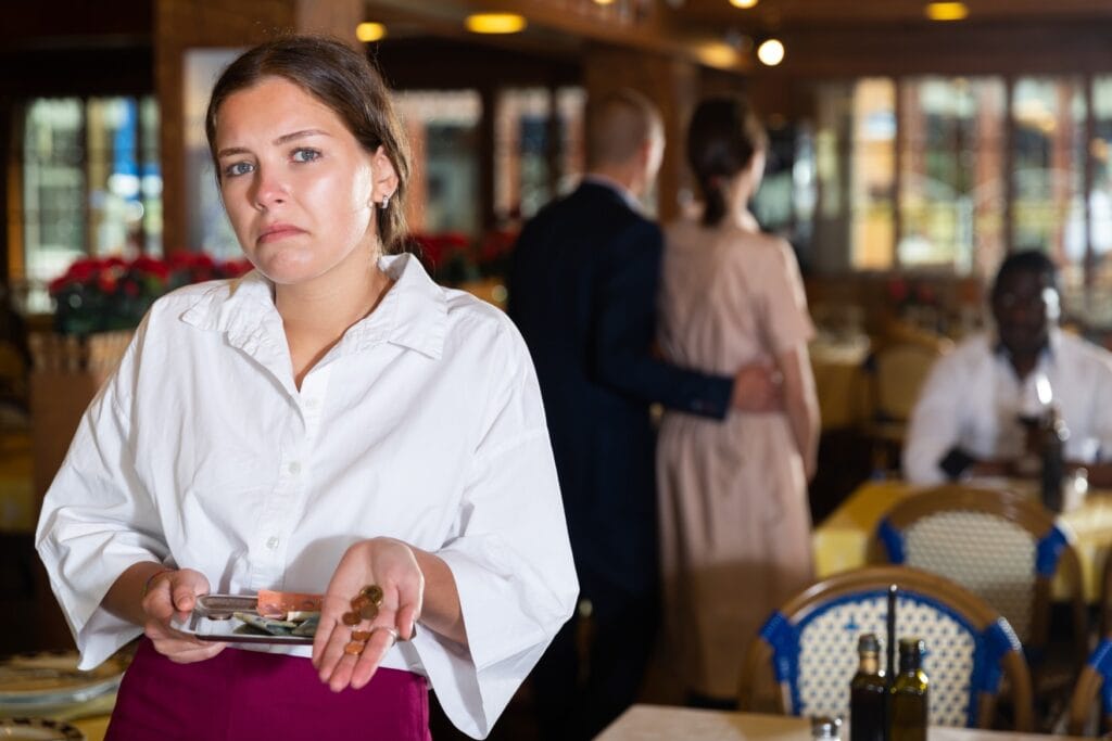 A waitress in a white shirt and a red shirt, holding a metal tablet with some euro bills on it and some low value cent coins in her other hand, looking disappointed into the camera. In the background a pair leaving the restaurant can be seen