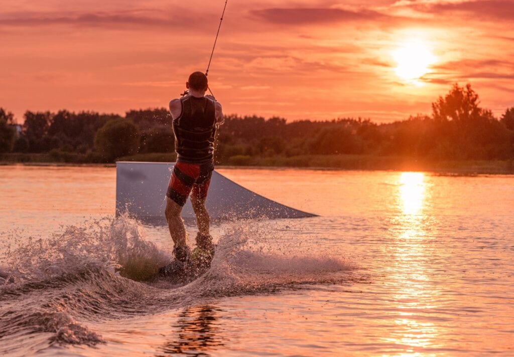 Eine Person mit Schwimmshorts und Weste hält sich an einem Seil fest und wird so auf einem Wakeboard vom Betrachter weg über einen See auf eine Rampe hin gezogen; im Hintergrund das bewaldete Seeufer, darüber taucht die untergehende Sonne das ganze Bild in ein rotes Licht