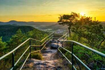 Ein felsiger Wanderweg, von Metallgeländern begrenzt, führt bis zu einer Sitzbank auf einer Hügelkuppe, zentral im Bild. Links und rechts vom Weg einige kleinere Bäume. Dahinter öffneet sich der Blick in eine Mittelgebirgslandschaft mit einer kleinen Stadt, alles beschienen von einem goldenen Sonnenuntergang