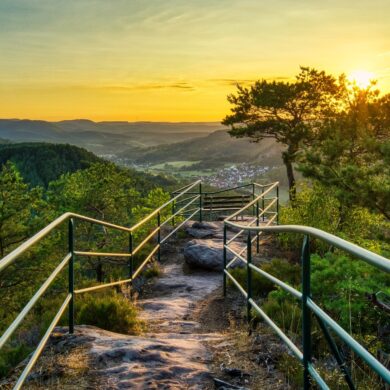 Ein felsiger Wanderweg, von Metallgeländern begrenzt, führt bis zu einer Sitzbank auf einer Hügelkuppe, zentral im Bild. Links und rechts vom Weg einige kleinere Bäume. Dahinter öffneet sich der Blick in eine Mittelgebirgslandschaft mit einer kleinen Stadt, alles beschienen von einem goldenen Sonnenuntergang