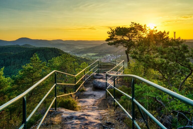 Ein felsiger Wanderweg, von Metallgeländern begrenzt, führt bis zu einer Sitzbank auf einer Hügelkuppe, zentral im Bild. Links und rechts vom Weg einige kleinere Bäume. Dahinter öffneet sich der Blick in eine Mittelgebirgslandschaft mit einer kleinen Stadt, alles beschienen von einem goldenen Sonnenuntergang