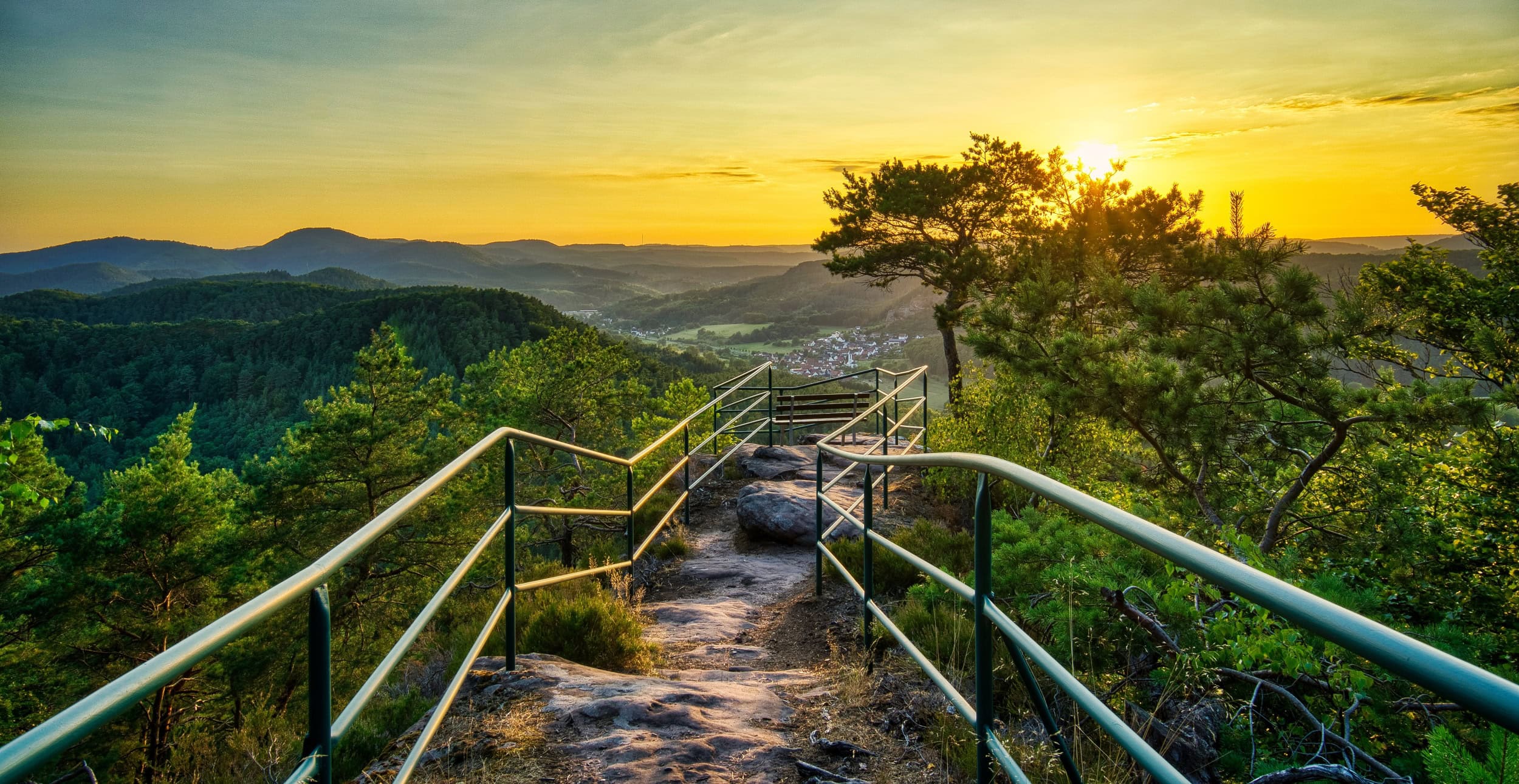 Ein felsiger Wanderweg, von Metallgeländern begrenzt, führt bis zu einer Sitzbank auf einer Hügelkuppe, zentral im Bild. Links und rechts vom Weg einige kleinere Bäume. Dahinter öffneet sich der Blick in eine Mittelgebirgslandschaft mit einer kleinen Stadt, alles beschienen von einem goldenen Sonnenuntergang