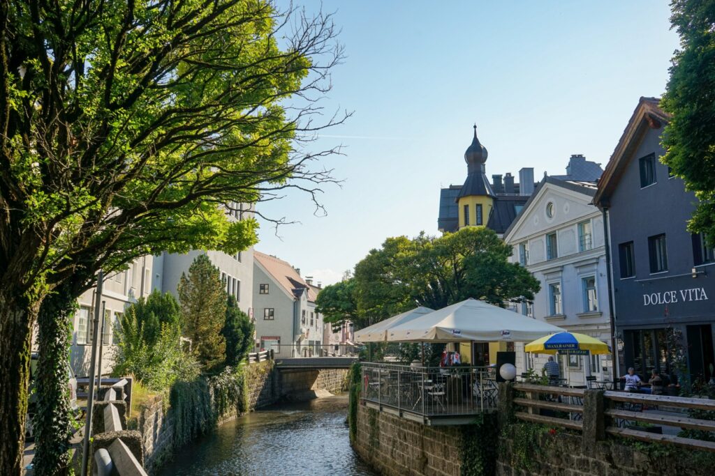 Ein kleiner, kanalisierter Fluss fließt längs durchs Bild, zu beiden Seiten reihen sich hübsche Häuser dicht an dicht. Auf der rechten Seite sind auf der Kanalstraße einige Sonnenschirme einer Außengastronomie aufgesspannt, an einer Häuserfassade prangt der Schriftzug "Dolce Vita".