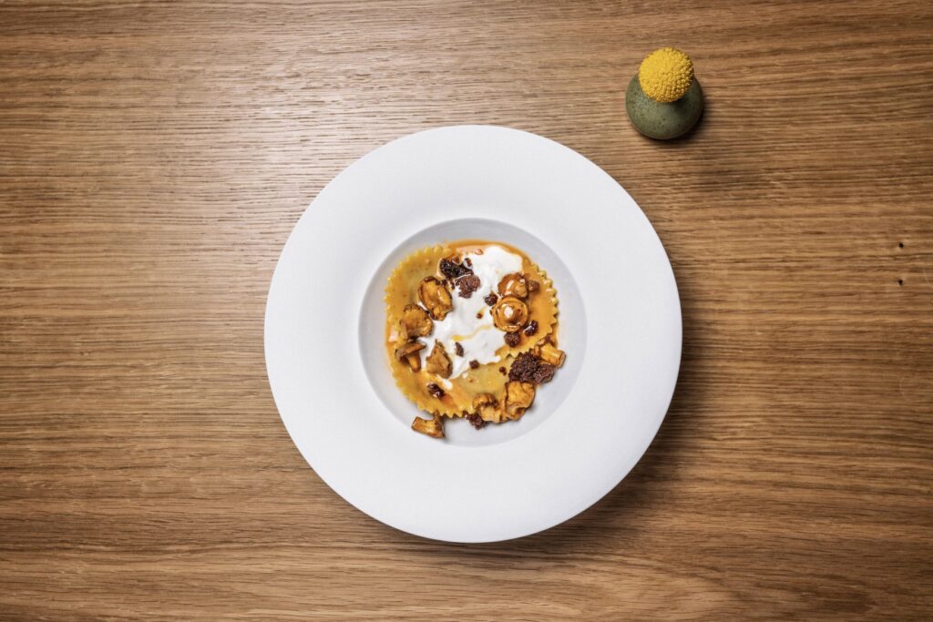 Three large ravioli are arranged on a white, deep plate with a few chanterelle mushrooms and a white Parmesan cream. The plate is photographed from directly above and stands in the center of a wooden table.