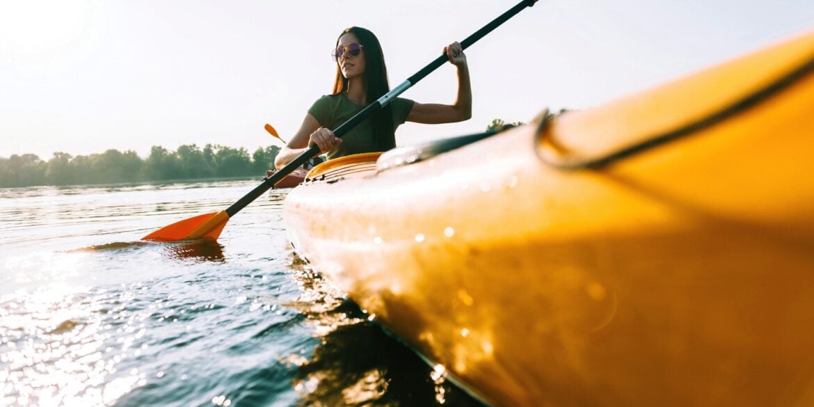 A woman wearing sunglasses and a green shirt dips a blade of her double paddle into the water from a bright yellow canoe. The canoe passes by the viewer on the right. In the background is a wooded shore.