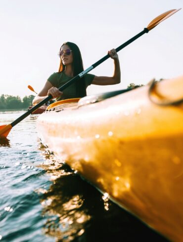 A woman wearing sunglasses and a green shirt dips a blade of her double paddle into the water from a bright yellow canoe. The canoe passes by the viewer on the right. In the background is a wooded shore.