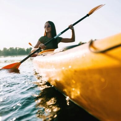 A woman wearing sunglasses and a green shirt dips a blade of her double paddle into the water from a bright yellow canoe. The canoe passes by the viewer on the right. In the background is a wooded shore.