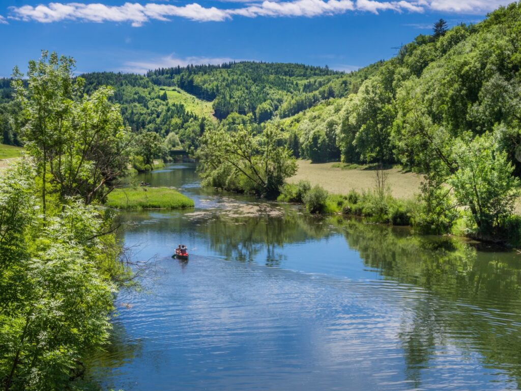 A wide river disappears behind a bend. There is light vegetation on the banks to the left and right. A small canoe with two people can be seen on the river. In the background, a wooded slope rises above a blue sky.