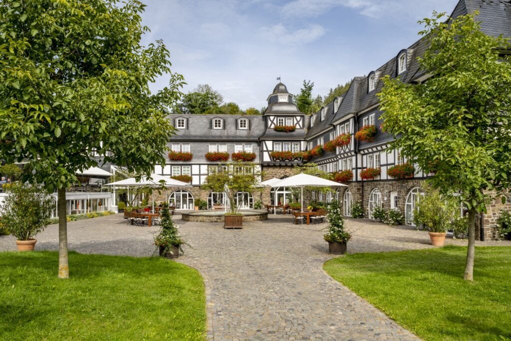 Um einen gepflasterten Hof legt sich dahinter und r echts davon ein dreigeschossiger Fachwerkhof mit schwarzem Schieferdach. An den Fenstern hängen Blumenkästen mit roten Blüten. Mittig auf dem Hof steht ein einfacher Springbrunnen, um den herum einige hölzerne Sitzgelegenheiten platziert wurden.