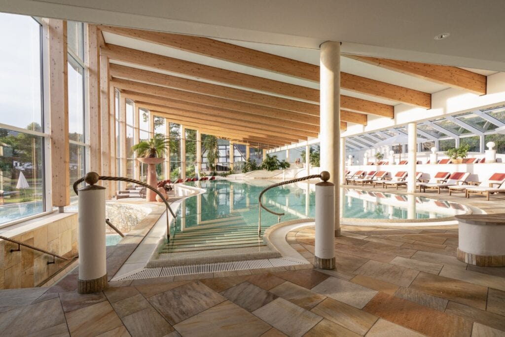 Von einem braun gefliesten Rand führt eine Treppe sanft in ein länglich-ovales Schwimmbecken in einer langen Halle mit Holzdecke. Rund um den Pool sind Liegen aufgestellt, durch eine lange Fensterwand auf der linken Seite fällt Tageslicht hinein.