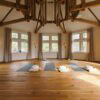 In einem achteckigen Raum mit weißen Wänden und großen Fenstern liegen drei Yogamatten mit Zubehör auf dem hölzernen Dieleboden. An der weißen Decke ist ein parallel zur Raumform laufendes Gebälk angebracht