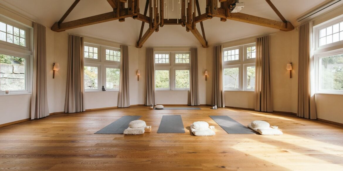 In einem achteckigen Raum mit weißen Wänden und großen Fenstern liegen drei Yogamatten mit Zubehör auf dem hölzernen Dieleboden. An der weißen Decke ist ein parallel zur Raumform laufendes Gebälk angebracht