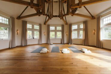 In einem achteckigen Raum mit weißen Wänden und großen Fenstern liegen drei Yogamatten mit Zubehör auf dem hölzernen Dieleboden. An der weißen Decke ist ein parallel zur Raumform laufendes Gebälk angebracht